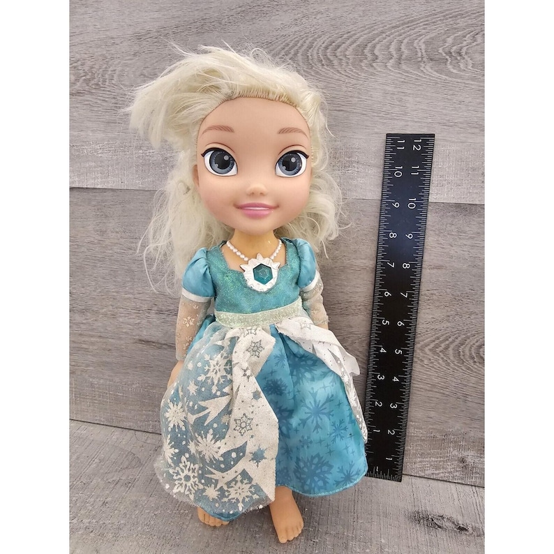 May include: A blonde doll wearing a blue dress with a snowflake pattern. The doll has blue eyes and is wearing a necklace with a blue gem. The doll is about 11 inches tall.