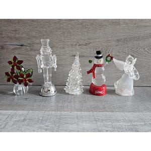 May include: Five clear glass Christmas ornaments with LED lights. The ornaments are a snowman, a Christmas tree, a nutcracker, an angel, and a poinsettia.