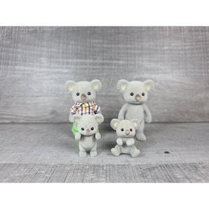 May include: Four gray koala figurines. Two are standing, one is sitting, and one is being held. One koala wears a plaid shirt. The koalas have small black eyes and tan noses. The background is a gray wooden surface.