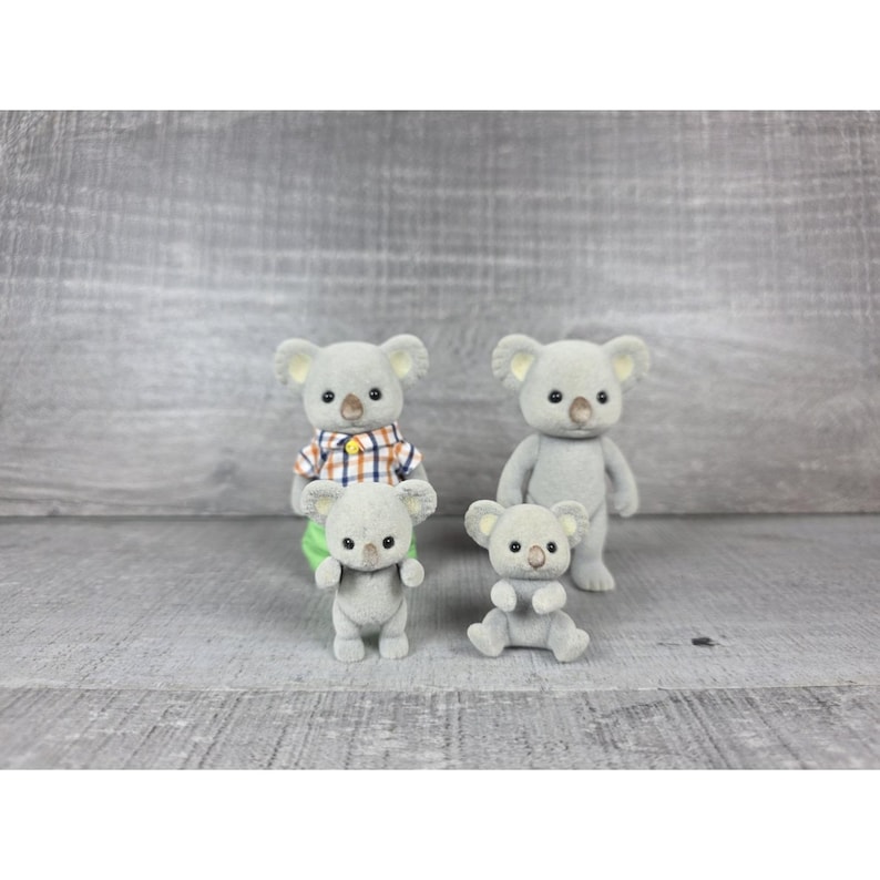 May include: Four gray koala figurines. Two are standing, one is sitting, and one is being held. One koala wears a plaid shirt. The koalas have small black eyes and tan noses. The background is a gray wooden surface.