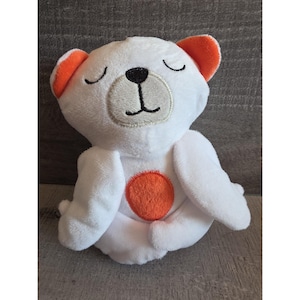 Könnte beinhalten: Ein weißes Plüschbär-Spielzeug mit orangefarbenen Akzenten an den Ohren und einem kreisförmigen Aufnäher auf der Brust. Der Bär hat geschlossene Augen und gestickte Gesichtszüge, darunter eine schwarze Nase und ein Mund.