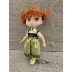 Puede incluir: Una mu&ntilde;eca peque&ntilde;a con cabello rojo, vestida con un vestido verde con un corpi&ntilde;o negro y un cuello blanco. La mu&ntilde;eca tiene ojos azules y una tez clara.