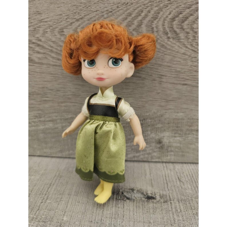 Puede incluir: Una mu&ntilde;eca peque&ntilde;a con cabello rojo, vestida con un vestido verde con un corpi&ntilde;o negro y un cuello blanco. La mu&ntilde;eca tiene ojos azules y una tez clara.