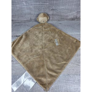 May include: A brown plush baby blanket with a monkey head attached. The blanket is square-shaped and has the word "LOVES" embroidered on it. The monkey has a tan face and ears. The blanket is made of soft material.