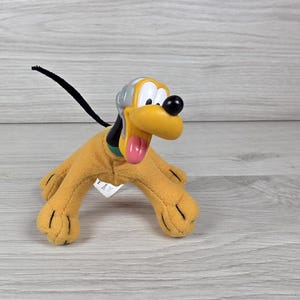 Pode incluir: Um peluche do Pluto com corpo amarelo, cauda preta e cabeça de plástico com um capacete cinzento. A personagem tem um nariz preto, uma língua cor-de-rosa e uma coleira verde. O brinquedo está de pé com quatro patas visíveis.