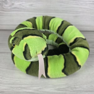 Puede incluir: Un juguete de serpiente de peluche enroscado en círculo, con una textura de tela suave y bandas alternas de color verde lima brillante, verde oliva oscuro y finas rayas negras. La serpiente tiene una lengua de fieltro bífida clara.