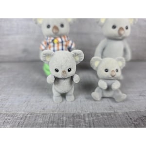 May include: Several small, gray koala figurines with black eyes and tan ears are arranged on a gray surface. One koala is standing, while another is seated. A koala in a plaid shirt is visible in the background.