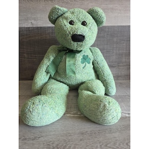 May include: A large, green teddy bear with a black nose and dark eyes. The bear has a green ribbon around its neck and a shamrock patch on its chest. The bear is sitting with its legs extended.