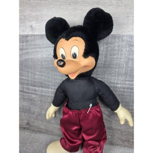 May include: Vintage Mickey Mouse plush doll with black fur ears and head, a black shirt, and maroon satin pants. The doll has a tan face, white gloves, and cream-colored feet. A white pin is attached to the shirt.