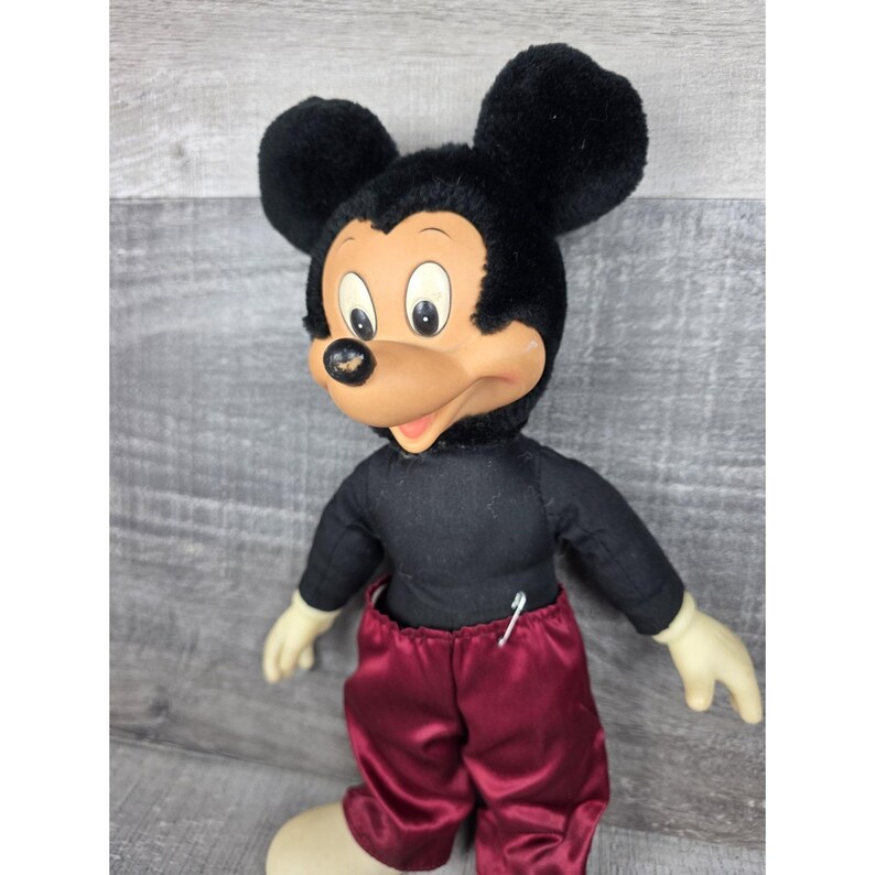 May include: Vintage Mickey Mouse plush doll with black fur ears and head, a black shirt, and maroon satin pants. The doll has a tan face, white gloves, and cream-colored feet. A white pin is attached to the shirt.