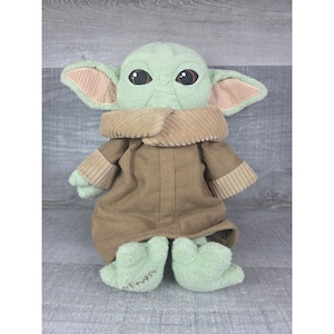 May include: A plush doll of a character with large eyes, green skin, and large ears. The doll wears a brown coat with a corduroy collar and cuffs. The doll's feet are green with embroidered details. The doll is approximately 12 inches tall.