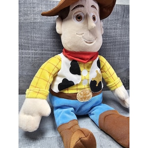 May include: A plush toy of the character Woody from the movie Toy Story. Woody is wearing a brown cowboy hat, a yellow plaid shirt, a white vest with black spots, blue pants, a red bandana, and brown boots. The toy has a smiling expression.