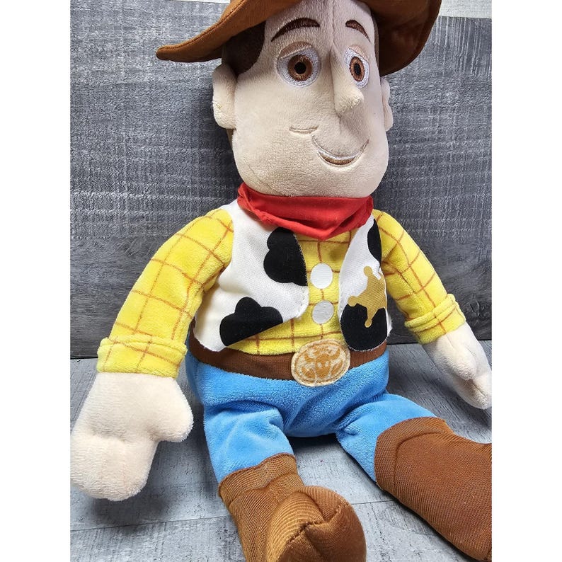 May include: A plush toy of the character Woody from the movie Toy Story. Woody is wearing a brown cowboy hat, a yellow plaid shirt, a white vest with black spots, blue pants, a red bandana, and brown boots. The toy has a smiling expression.