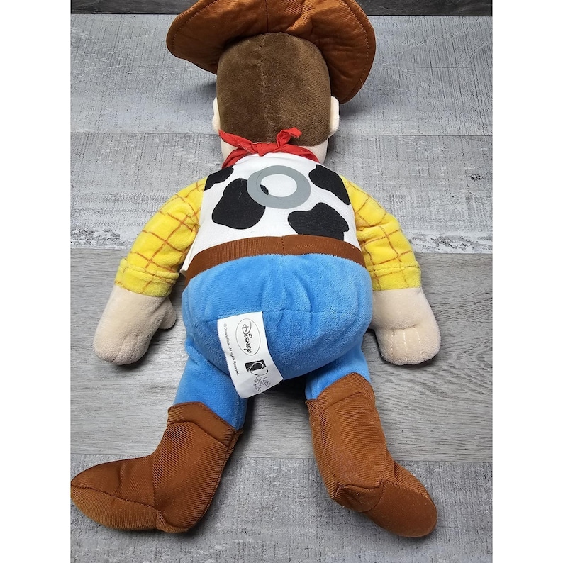 May include: A plush toy of Woody from Toy Story, viewed from the back. The doll features a brown cowboy hat, a yellow plaid shirt, blue pants, and brown boots. The vest has a cowhide pattern. A red scarf is around the neck.