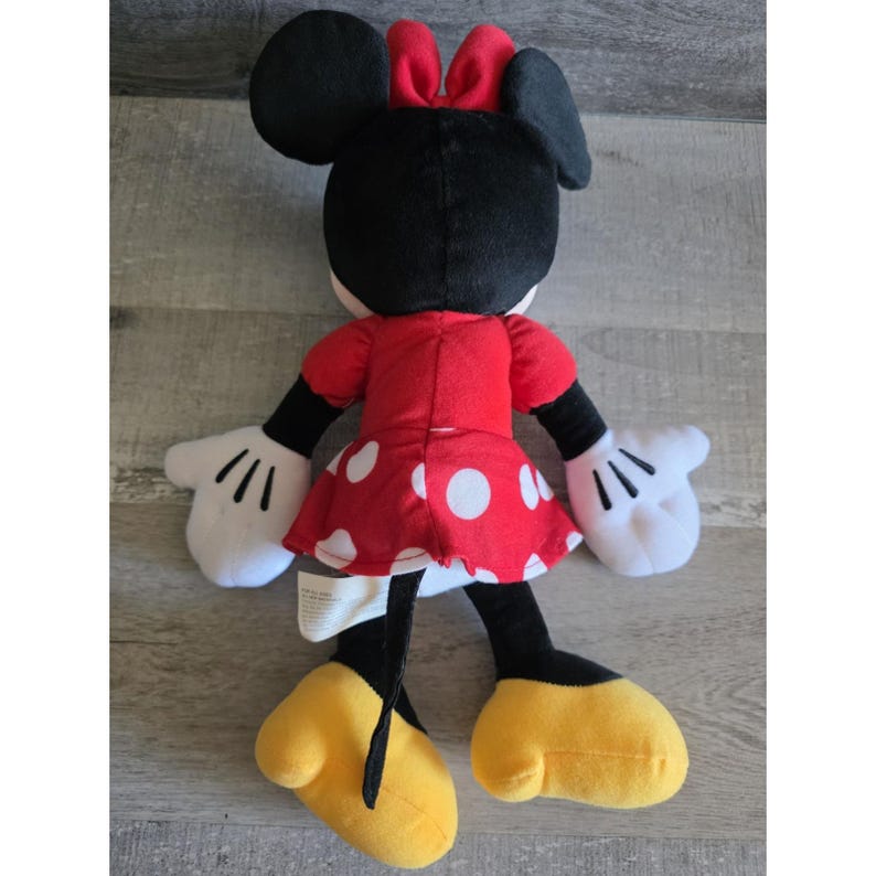 May include: A plush Minnie Mouse doll, viewed from the back, features a red dress with white polka dots, a red bow, and black ears. The doll has white gloves and yellow shoes. The doll is made of soft fabric and is designed for children.