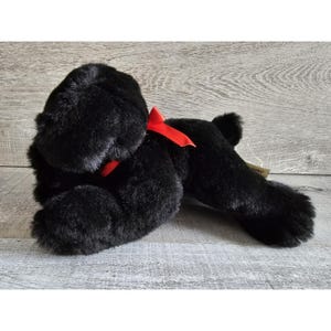 Puede incluir: Perro de peluche negro con un lazo rojo. El perro está acostado boca abajo con la cabeza girada hacia un lado.