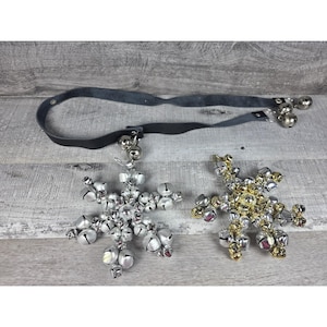 May include: Two snowflake-shaped ornaments constructed from numerous small jingle bells. One ornament is silver-toned, and the other is a mix of gold and silver tones. A long, thin black leather strap with additional jingle bells is attached to the silver snowflake.