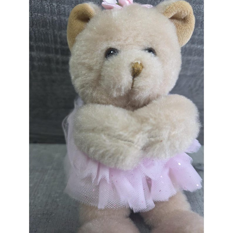 Pu&ograve; includere: Un orsacchiotto di peluche con la pelliccia marrone chiaro, che indossa un tut&ugrave; rosa e un fiore rosa sulla testa. Le braccia dell'orso sono giunte. Il tut&ugrave; &egrave; fatto di un tessuto trasparente e scintillante.