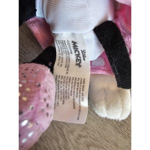 May include: Pink and white plush Mickey Mouse toy with black ears and a pink polka dot bow. The tag reads "Disney Mickey" and includes care instructions and licensing information.