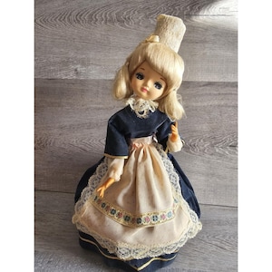 May include: A vintage doll wearing a blue velvet dress with a white lace trim and a white apron. The doll has blonde hair and is wearing a white hat.