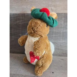 May include: A plush brown bunny wearing a green and red hat, holding a red heart-shaped object. The bunny has a cream-colored belly and a white fluffy tail. The toy is set against a wooden background.