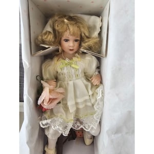 Puede incluir: Una muñeca de porcelana con cabello rubio y un vestido blanco con ribete de encaje. Lleva una rosa rosa y una cinta verde alrededor del cuello.