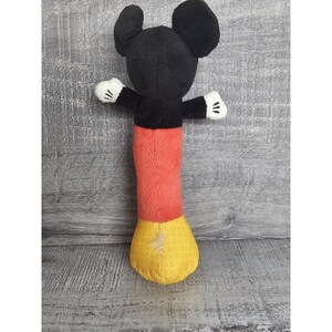 May include: A plush Mickey Mouse toy with black ears, a black torso, red trousers, and yellow patterned feet. The toy has white gloves and outstretched arms.