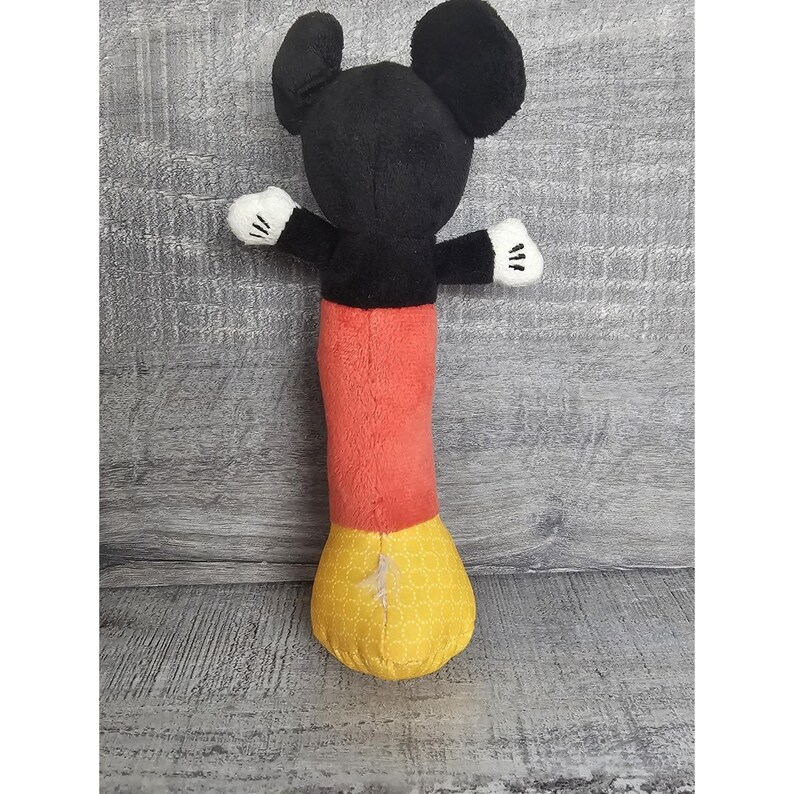 May include: A plush Mickey Mouse toy with black ears, a black torso, red trousers, and yellow patterned feet. The toy has white gloves and outstretched arms.