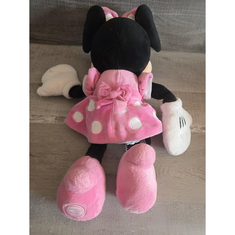 May include: A plush Minnie Mouse doll, seen from the back, has a pink dress with white polka dots and a matching bow. The doll has black ears, arms, and legs, with white gloves and pink shoes. The doll is made of soft, plush material.