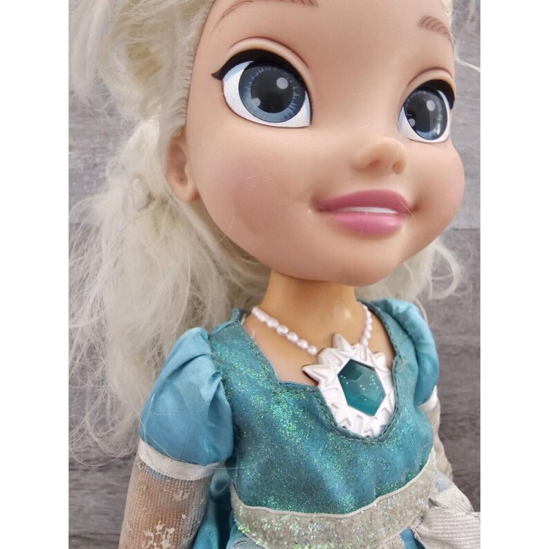May include: A blonde doll wearing a blue and silver dress with a snowflake pendant necklace. The doll has blue eyes and a pink smile.