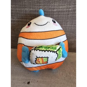 May include: A plush toy shaped like a sushi roll with a smiling face. The toy is white with orange stripes and has blue arms. The toy is holding a green and yellow sushi roll.