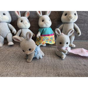 Puede incluir: Una colecci&oacute;n de figuras de conejos de juguete grises flocados. Algunos conejos llevan vestidos y lazos. Los conejos tienen orejas largas y ojos negros. Los juguetes est&aacute;n colocados sobre una superficie gris.