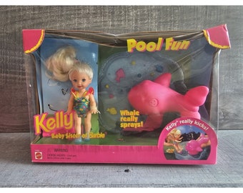 Puede incluir: Un juguete de ballena de plástico rosa con una función de pulverización de agua. El juguete se empaqueta con una muñeca y un juego de piscina. La caja presenta el texto "Pool Fun" y "Kelly really kicks!"