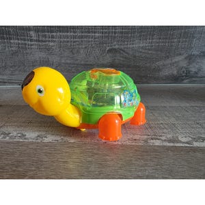 May include: A brightly coloured toy turtle in yellow, orange, and green. The turtle has a clear green shell with a graphic, orange feet, and a brown spot on its head. A fun toy for children.