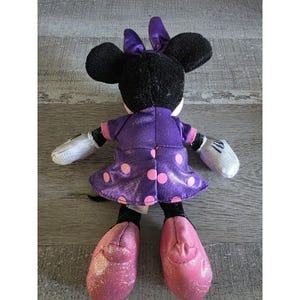 Op de afbeelding: Een pluche Minnie Mouse pop, gezien van achteren, met een paarse jurk met roze stippen, een bijpassende strik en roze glitter schoenen. De pop heeft zwarte oren en witte handschoenen.