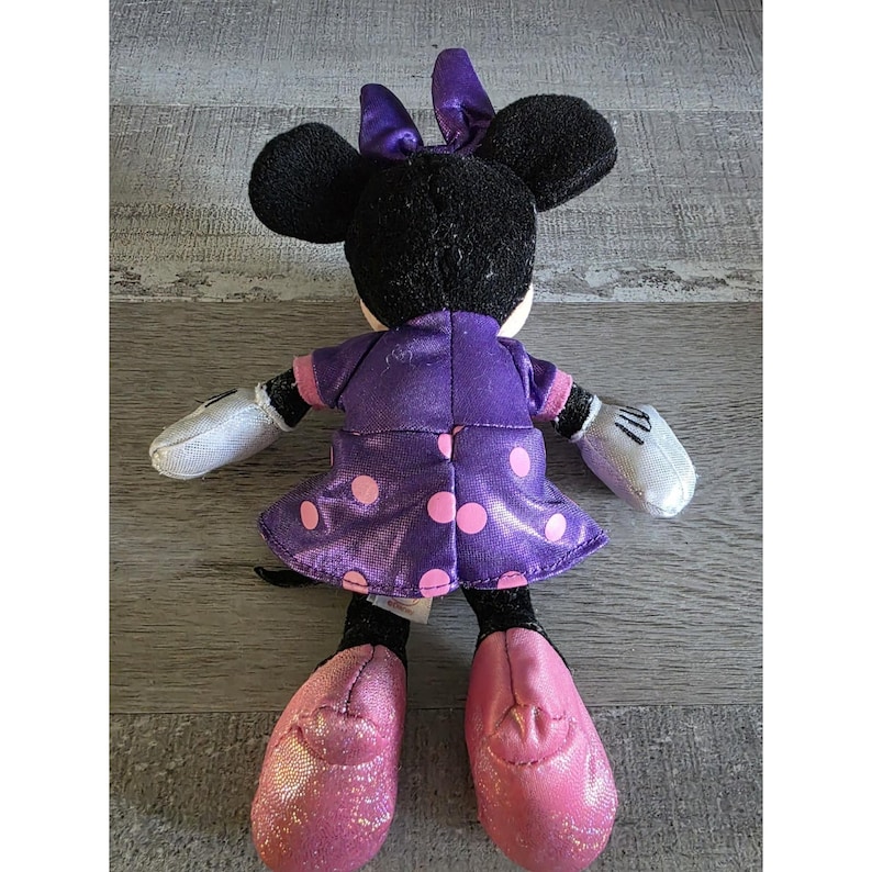 Op de afbeelding: Een pluche Minnie Mouse pop, gezien van achteren, met een paarse jurk met roze stippen, een bijpassende strik en roze glitter schoenen. De pop heeft zwarte oren en witte handschoenen.