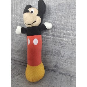 May include: A Mickey Mouse soft toy with a black head and ears, a beige face, and white hands. The body is red with two white circles, and the feet are yellow with a geometric pattern. The toy is upright with one arm raised.