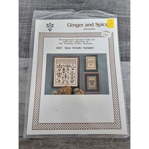 Pode incluir: Um padrão de bordado de ponto cruz em papel branco com um sampler do Reading Public Museum. O sampler é intitulado "1827 Sara Schultz Sampler" e inclui uma variedade de motivos florais e geométricos. O padrão é de Ginger and Spice.