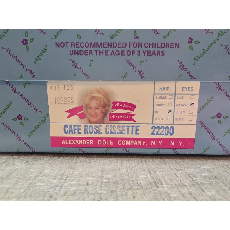 May include: A close-up of a box for a Madame Alexander doll. The box is light blue with a floral pattern and the words "Madame Alexander" printed on it. The label reads "Cafe Rose Cissette 22200".