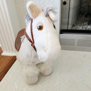 Peut inclure: Une peluche en forme de cheval blanc à la texture douce, dotée d'une selle en similicuir marron, d'une bride marron, ainsi que d'une crinière et d'une queue gris-blanc. Le cheval a de grands yeux marron ronds et des taches gris clair sur le corps.