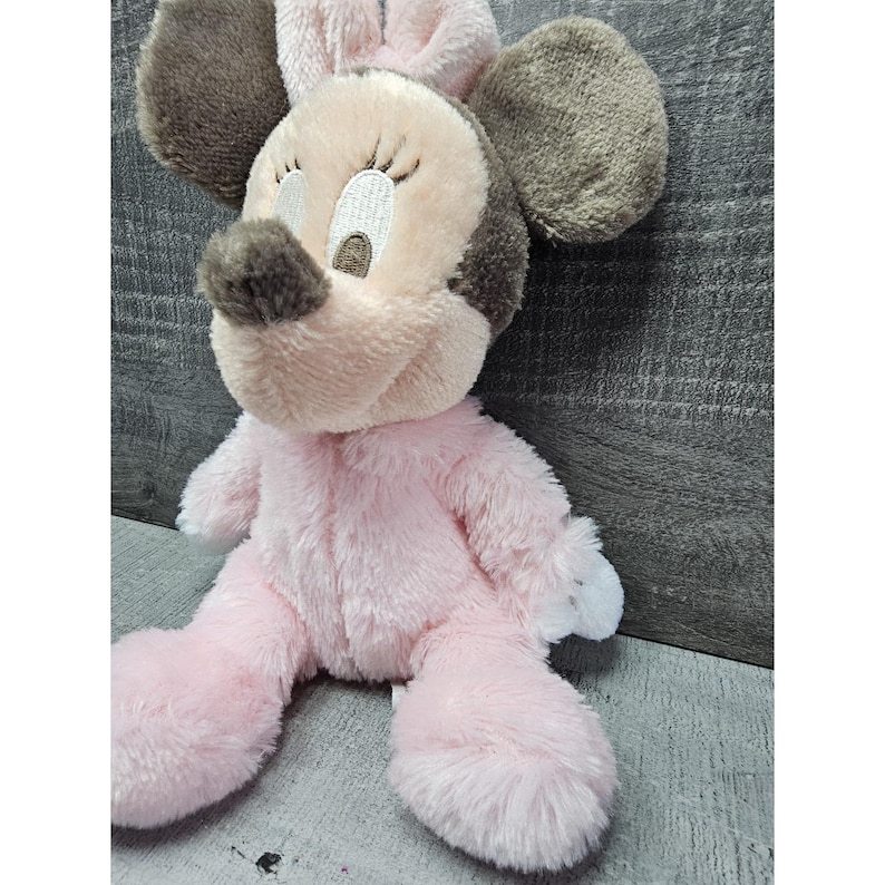 May include: A plush Minnie Mouse doll in a seated position. The doll features a pink body, a light pink face, and gray ears. The eyes are embroidered with black eyelashes, and the nose is brown. The doll is a soft, cuddly toy.