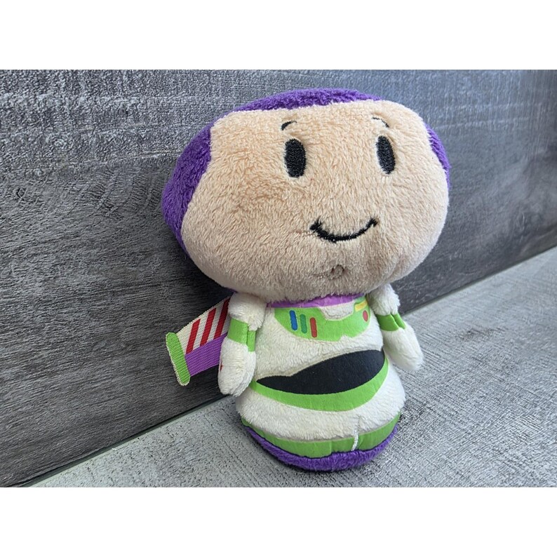 May include: A plush Buzz Lightyear toy with a purple head, tan face, and a smiling expression. The toy is wearing a white and green spacesuit with a green stripe and a red, white, and purple wing. The toy is approximately 6 inches tall.