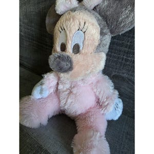 May include: A plush Minnie Mouse doll in pink, gray, and white. The doll has large, embroidered eyes, a gray nose, and a pink body. The doll's ears are gray, and the feet and hands are white.
