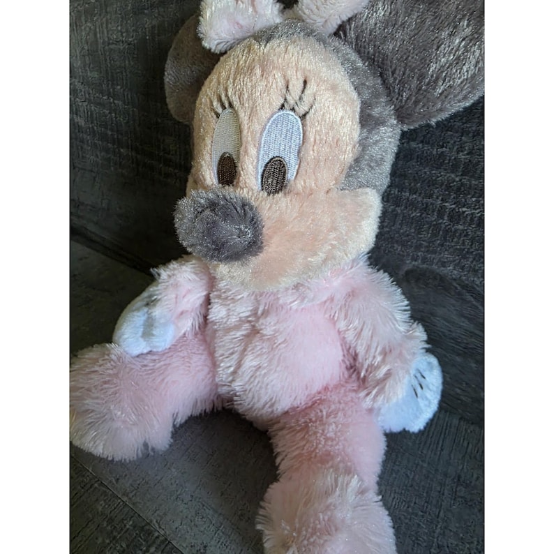 May include: A plush Minnie Mouse doll in pink, gray, and white. The doll has large, embroidered eyes, a gray nose, and a pink body. The doll's ears are gray, and the feet and hands are white.