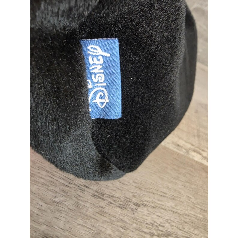 May include: Close-up of a black plush toy with a blue tag that reads "Disney" in white script. The plush toy has a soft, fuzzy texture. The tag is rectangular and has rounded corners.