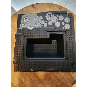 Könnte beinhalten: Schwarze Lego-Aufbewahrungsbox mit einem grauen und weißen abstrakten Design auf der Oberseite. Die Box hat eine rechteckige Öffnung an der Vorderseite.