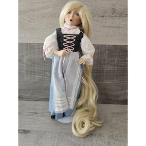 Puede incluir: Una muñeca de porcelana con el pelo largo y rubio que lleva una blusa blanca, un chaleco negro con cordones rosas y una falda azul con ribete de encaje blanco.