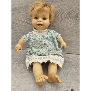 May include: A vintage baby doll with blonde hair, blue eyes, and a wide smile. The doll is wearing a white dress with blue floral print and white lace trim.