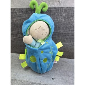 May include: A plush baby toy in a blue hooded outfit with green accents. The toy has a light beige face with closed eyes and a small smile. The outfit has green circles and yellow ribbons. The text "by Bug" is visible.