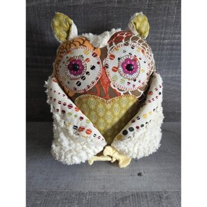 May include: A handmade owl-shaped plush toy with a patchwork design. The owl features large, circular eyes with pink centers, a brown body, and white fluffy accents. The wings and ears are adorned with patterned fabrics in various colors.